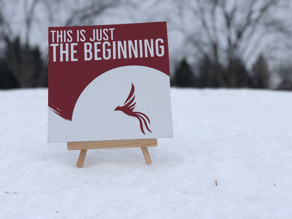 Photo of a canvas sign that says "This is just the beginning" with a red phoenix in the bottom. The background is a winter scene.