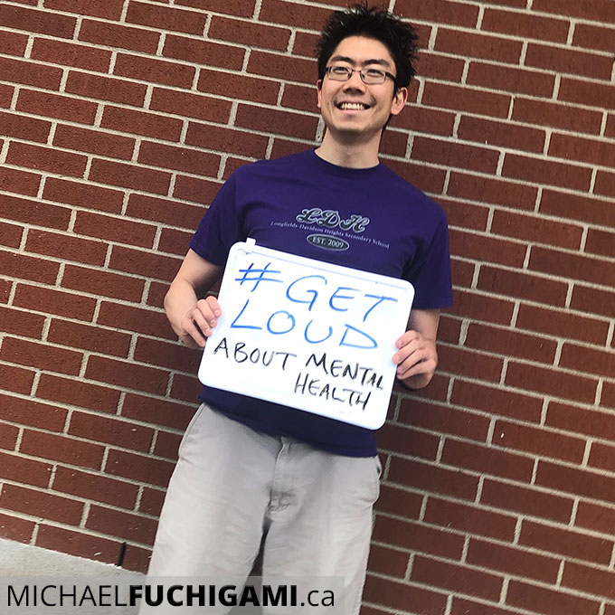 Photo of Mike Fuchigami holding up a sign that says #GetLoud about mental health. 